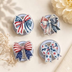May include: Four circular phone grips featuring decorative bow attachments. Each bow displays a patriotic theme with red, white, and blue colors, incorporating star and stripe patterns. The grips are arranged in a two-by-two grid configuration.