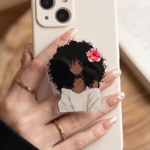 May include: A clear phone grip featuring a black-haired woman with a flower in her hair. The woman is wearing a white top and a gold necklace. The phone case is off-white.