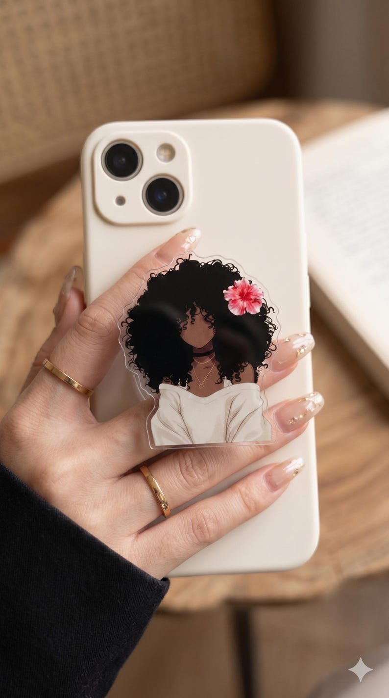 May include: A clear phone grip featuring a black-haired woman with a flower in her hair. The woman is wearing a white top and a gold necklace. The phone case is off-white.