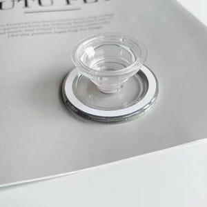 May include: A clear, transparent, plastic funnel-shaped object with a circular base and a white rim. The object is on a light grey surface. The funnel has a small opening at the bottom.