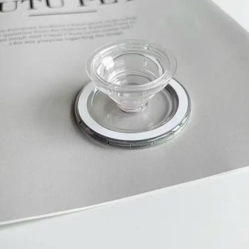 May include: A clear, transparent, plastic funnel-shaped object with a circular base and a white rim. The object is on a light grey surface. The funnel has a small opening at the bottom.