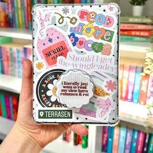 Könnte beinhalten: Eine transparente Handyhülle mit bunten Aufklebern mit Texten wie "Read More Books" und "Should I get the wingleader". Die Hülle hat einen Pop-Socket mit dem Text "i literally just want to read my slow burn romance & roi".
