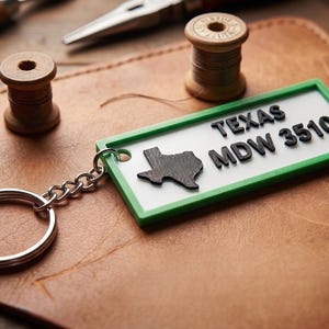 May include: A rectangular keychain with a green border and the text "TEXAS MDW 3510" in black. The keychain features a black silhouette of the state of Texas. It is attached to a silver key ring. The background includes leather and wooden spools.