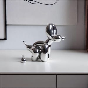 May include: A reflective silver-coloured sculpture shaped like a balloon animal dog in a crouching position, accompanied by a small, separate silver-coloured spherical object placed on the surface near the rear of the dog figure.