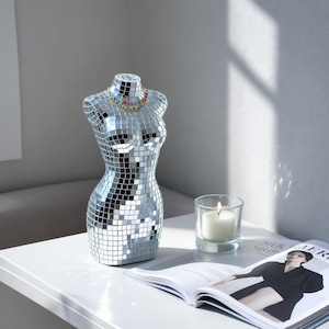 May include: A torso-shaped sculpture covered in small, reflective silver mirror tiles, creating a disco ball effect. The sculpture features a multi-colored beaded necklace around the neck area, with a clear glass candle holder nearby.