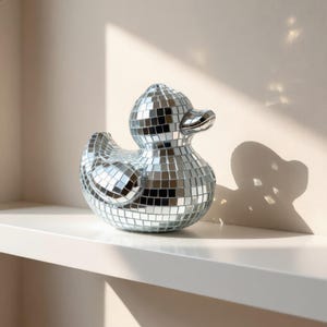May include: A decorative silver duck figurine covered in small, square mirrored tiles. The duck is positioned on a white shelf against a neutral background. The duck's beak and eyes are visible.