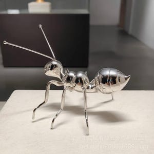 May include: A polished silver-colored metal ant figurine with long antennae and six slender legs. The surface is highly reflective, showing a smooth, segmented body. The text "东莞市东城昕毅五金模具加工厂 heliwujin2008.1688.com" is visible at the bottom.