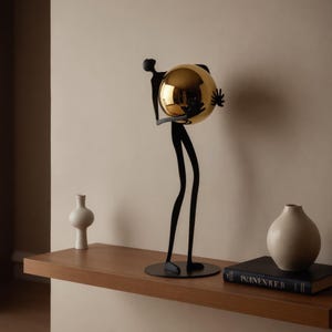 Pode incluir: Uma escultura alta e esguia de metal preto que representa uma figura estilizada sobre uma base circular, segurando uma grande esfera de cor dourada polida nas mãos. A figura tem membros alongados e um acabamento preto mate suave.