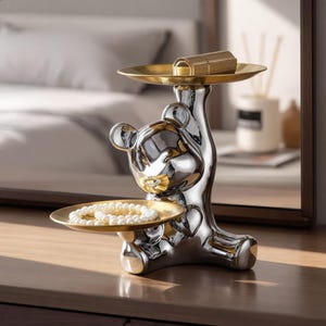 May include: A silver-toned bear figurine serving as a decorative stand, featuring two circular gold-toned trays. The top tray holds a rectangular gold object, while the lower tray holds a strand of white pearls. The bear has a reflective finish.
