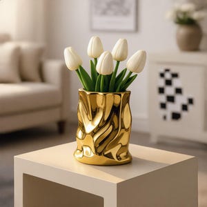 May include: A metallic gold vase with a crinkled, irregular surface texture containing a bouquet of five cream-colored tulip flowers with green stems and leaves. The vase has a reflective finish and a cylindrical shape with a wavy, uneven rim.