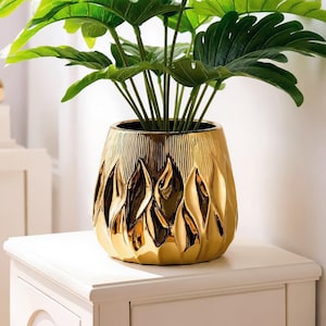 May include: A shiny gold ceramic planter with a textured, flame-like embossed pattern on the exterior. The planter holds a lush, green artificial plant with large, broad, split-leaf foliage that extends upwards from the center of the vessel.