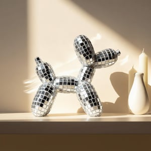 May include: A decorative dog sculpture made of mirrored tiles, reflecting light. The dog is in a playful pose, with a white vase and candles in the background. The sculpture is a unique home decor item.