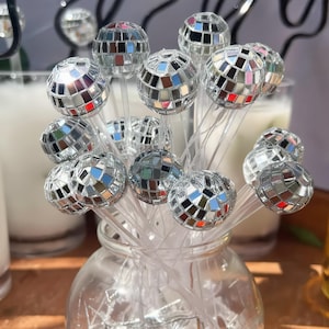 Peut inclure: Un ensemble de mélangeurs à boisson en plastique transparent, chacun surmonté d'une petite boule à facettes sphérique recouverte de minuscules carreaux de miroir argentés réfléchissants. Les mélangeurs sont regroupés dans un récipient en verre.