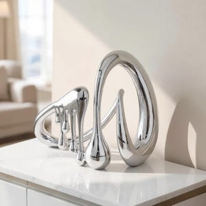 May include: A silver-coloured abstract sculpture with a polished, reflective finish. The object features fluid, curved lines and multiple teardrop-shaped elements that appear to be dripping downward, creating a dynamic and interconnected metallic form.