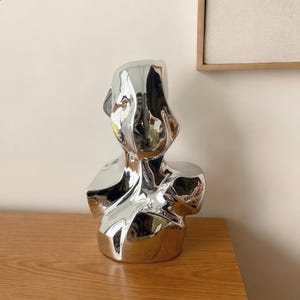 Puede incluir: Una escultura de color plateado reflectante con forma de busto humano. La superficie es lisa y está muy pulida, creando reflejos distorsionados similares a un espejo del entorno circundante en su forma irregular, fluida y abstracta.
