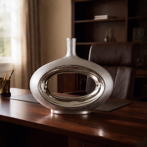 May include: A silver-toned vase with a wide, flattened oval body and a central hollow opening. The exterior surface features a textured, matte finish, while the inner rim of the central cutout has a smooth, reflective, polished metallic surface.