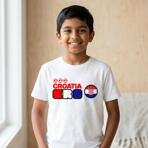 Puede incluir: Una camiseta blanca de manga corta con un dise&ntilde;o gr&aacute;fico que incluye tres balones de f&uacute;tbol rojos, la palabra CROATIA en rojo, las letras CRO en rojo, blanco y azul, y un emblema circular de la bandera croata con un patr&oacute;n de cuadros rojo y blanco.