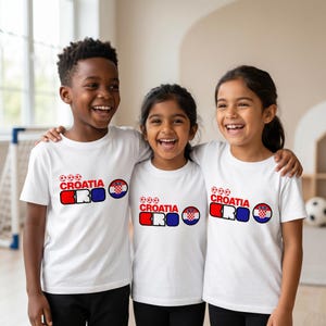 Puede incluir: Tres camisetas blancas de manga corta con un dise&ntilde;o gr&aacute;fico que incluye tres balones de f&uacute;tbol rojos, la palabra CROATIA en rojo, las letras CRO en rojo, blanco y azul, y un emblema circular de la bandera croata en el pecho de cada prenda.