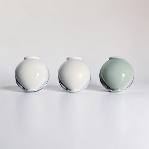 May include: Three ceramic vases in a row. Two are white, and one is a pale green. Each vase has a rounded body and a small, cylindrical neck. The vases have a clear, curved base that wraps around the bottom of the vase.