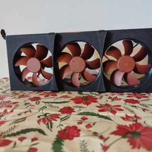 Pode incluir: Um conjunto de ventoinhas de computador pretas com três lâminas castanhas. As ventoinhas estão alojadas numa estrutura retangular preta. O conjunto está sobre uma superfície com padrão floral.