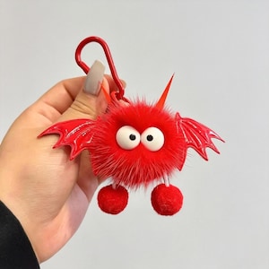 Puede incluir: Un llavero de monstruo rojo y peludo con ojos blancos, alas rojas, cuernos y pies de pompón. El llavero tiene un cierre de metal rojo. El monstruo tiene un diseño de dibujos animados.