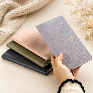 May include: A hand holds a light gray, textured To-Do List journal with gold lettering. Other journals in pink, olive green, and navy blue are stacked below. The journals have a leather-like appearance and include a ribbon bookmark.