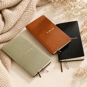 May include: Three leather-bound notebooks in shades of sage green, brown, and black. Each notebook has a textured cover and a ribbon bookmark. The brown notebook is labeled "To-Do List", and the black notebook is labeled "Do List".