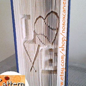 Könnte beinhalten: Eine Buchkunstskulptur des Wortes "LOVE" mit einem Herzen in der Mitte. Das Buch ist gefaltet und geformt, um die Buchstaben und das Herz zu bilden. Das Buch ist blau am Rücken und die Seiten sind weiß. Der Text "Pattern Only" ist in der linken unteren Ecke des Bildes sichtbar. Der Text "www.etsy.com/shop/nosoxcanves" ist auf der rechten Seite des Buches sichtbar.
