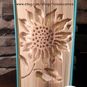 Könnte beinhalten: Eine Buchkunstskulptur einer Sonnenblume, gefaltet aus den Seiten eines Buches. Das Buch ist in blaugrün gebunden und die Skulptur steht auf einer Holzoberfläche. Der Text "www.etsy.com/shop/nosoxcarves" ist auf das Buch gedruckt. Der Text "Pattern Only" ist auf einen weißen Kreis mit buntem Hintergrund gedruckt.