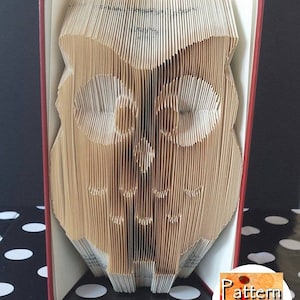 Peut inclure: Un livre dont les pages sont pliées en forme de hibou. Le livre est rouge et le hibou est brun. Le livre est posé sur une surface à pois noirs et blancs. Le texte "Pattern Only" est visible dans le coin inférieur droit de l'image.