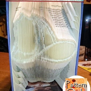 May include: A book with pages folded into a 3D shape resembling a heart. The book is bound in blue and has a silver ribbon around it. The text "Pattern Only" is visible on a circular sticker in the lower right corner of the image.