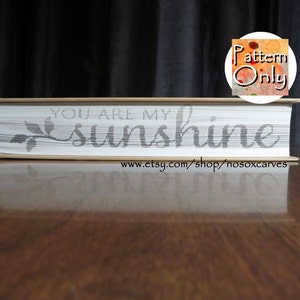 以下が含まれることがあります： 白い本の表紙に「You are my sunshine」という文字が彫られています。本は木製の表面に置かれています。画像の右上隅には円の中に「Pattern Only」という文字が見えます。
