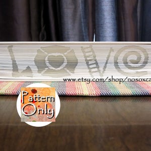 May include: A white book with a pattern carved into the pages. The pattern includes a heart, a ladder, a key, and a spiral. The book is sitting on a striped fabric.