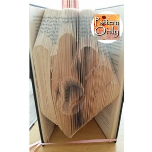 May include: A book with folded pages to create a 3D design of a heart. The book is open and the pages are folded to create a heart shape. The book is bound in black and has a pink ribbon tied around it. The text "Pattern Only" is visible on a sticker on the book.