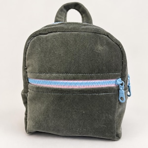 May include: A small dark olive green velvet backpack featuring a top handle and a front pocket. The front pocket is secured with a light blue zip that has a pink fabric trim. The backpack has a rounded shape and a soft, textured fabric finish.