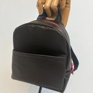 Puede incluir: Una mochila pequeña de color marrón oscuro con textura lisa, que cuenta con un bolsillo frontal y una cremallera de color rosa brillante en el compartimento superior. Incluye un asa superior y correas ajustables para los hombros.