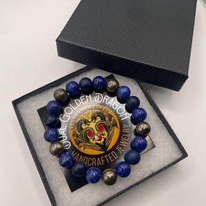 May include: A beaded bracelet featuring alternating dark blue and metallic grey spherical beads, presented in a black textured gift box. The bracelet rests on a circular card with a dragon illustration and the text Dual Golden Dragon Handcrafted Jewelry.