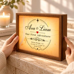May include: A rectangular wooden light box featuring a backlit map design. The text reads Ava & Liam, Our First Adventure, Baton Rouge, LA, 30.4344 N, 91.0394 W, and 08.08.2026. A small red heart icon sits between the names.