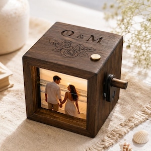 May include: A dark brown wooden music box with a square frame featuring a photograph of a couple on a beach. The top surface is engraved with the letters O & M and a rose illustration, and a small wooden crank handle is attached to the side.