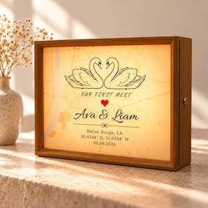 Peut inclure: Une boîte lumineuse rectangulaire en bois avec un fond de carte et deux cygnes se faisant face. Le texte indique : OUR FIRST MEET, un cœur rouge, Ava & Liam, Baton Rouge, LA, 30.4344 N, 91.0394 W, et la date 08.08.2026.