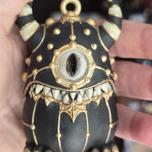 May include: A small, black, rounded creature figurine with a single central eye, two curved horns, and sharp white teeth. The figure is decorated with gold-colored accents, including a circular frame around the eye, studs, and vertical lines.