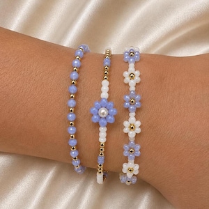Può includere: Un set di tre braccialetti di perline con perline sfaccettate azzurre, piccole sfere metalliche dorate, perline rotonde bianche e motivi a forma di fiore realizzati con perline azzurre e bianche con accenti centrali bianchi o dorati.