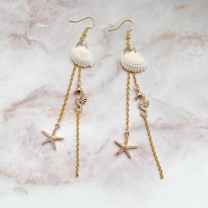 Könnte beinhalten: Ohrringe zum Aufhängen mit Strandmotiv. Jeder Ohrring hat eine weiße Muschel, einen goldenen Seepferdchen-Anhänger und einen goldenen Seestern-Anhänger. Die Ohrringe haben goldfarbene Ohrhaken und goldfarbene Ketten.