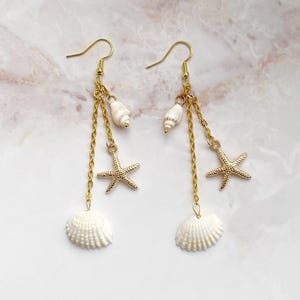Könnte beinhalten: Goldfarbene Hängeohrringe mit Muschel-, Seestern- und kleinem Muscheldesign. Die Ohrringe haben eine Kette und einen Hakenverschluss. Die Muscheln sind weiß und die Seesterne sind goldfarben.