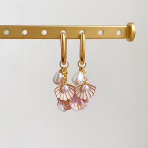 Könnte beinhalten: Goldene Creolen mit Muschel- und Perlenanhängern. Die Ohrringe haben schillernde rosa Muschel-Anhänger und weiße, tropfenförmige Perlen. Sie hängen an einer goldenen Stange.