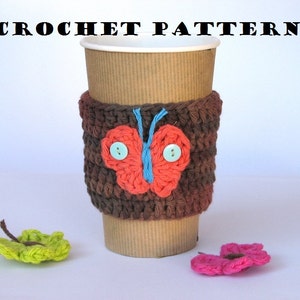 Könnte beinhalten: Eine braune gehäkelte Kaffeetassen-Hülle mit einem roten Schmetterlingsdesign und blauen Knöpfen. Die Hülle befindet sich auf einem braunen Pappbecher. Auf der weißen Oberfläche befinden sich zwei gehäkelte Schmetterlinge.