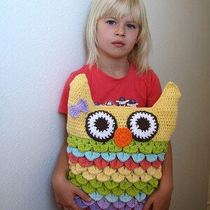 Puede incluir: Una almohada de b&uacute;ho de crochet con un esquema de color amarillo, verde, naranja y morado. El b&uacute;ho tiene grandes ojos blancos con pupilas marrones y una nariz naranja brillante. La almohada la sostiene un ni&ntilde;o que lleva una camisa roja y pantalones cortos de mezclilla azules.