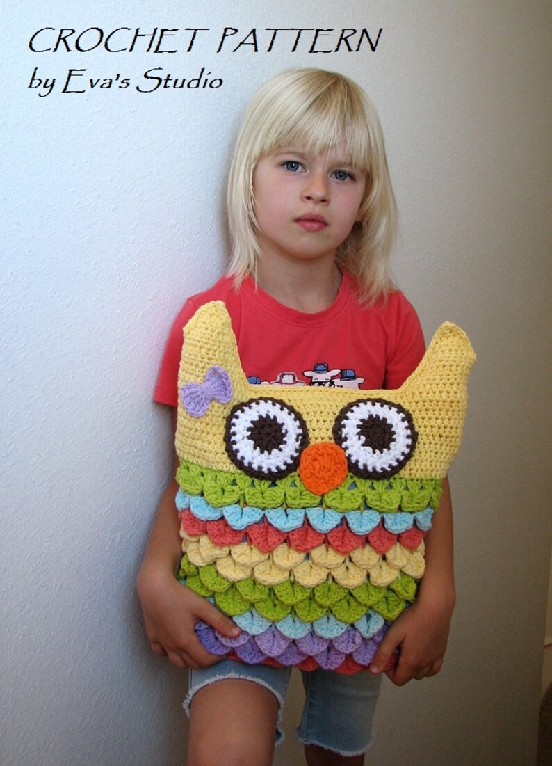 Puede incluir: Una almohada de b&uacute;ho de crochet con un esquema de color amarillo, verde, naranja y morado. El b&uacute;ho tiene grandes ojos blancos con pupilas marrones y una nariz naranja brillante. La almohada la sostiene un ni&ntilde;o que lleva una camisa roja y pantalones cortos de mezclilla azules.