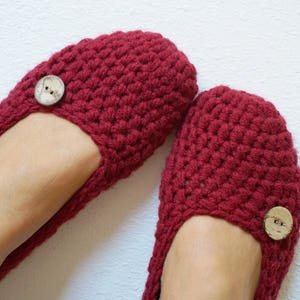 Könnte beinhalten: Ein Paar weinrote Häkelslipper mit Knopfdetail. Die handgefertigten Slipper wirken weich und bequem. Der Knopf ist hellbraun. Die Slipper werden auf weißem Hintergrund gezeigt. Ideal für den Innenbereich.