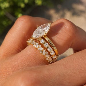 May include: A gold ring set consisting of three stacked bands. The top band features a large marquise-cut center stone. The middle band is set with round stones, and the bottom band displays a pattern of alternating baguette and marquise-cut stones.